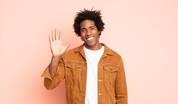 Joven afro negro sonriendo feliz y alegremente, saludando con la mano, dándole la bienvenida y saludándolo