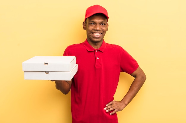 Joven afro negro sonriendo felizmente con una mano en la cadera y confiado concepto de repartidor de pizza