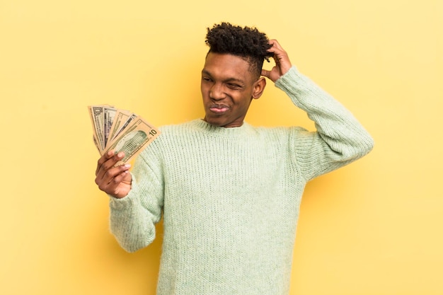 Joven afro negro sonriendo felizmente y soñando despierto o dudando del concepto de billetes de banco en dólares