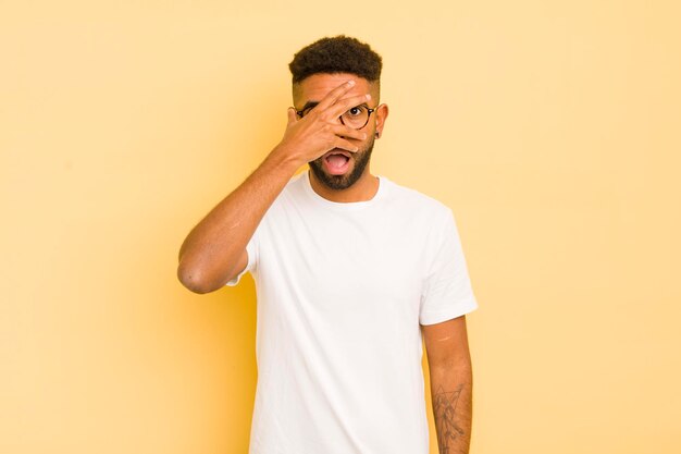 Joven afro que parece sorprendido, asustado o aterrorizado cubriendo la cara con la mano y mirando entre los dedos