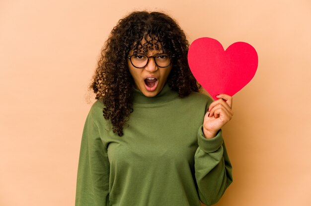 Joven afroamericana afro sosteniendo un corazón de San Valentín gritando muy enojado y agresivo.