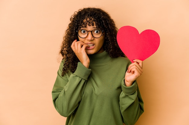 Joven afroamericana afro sosteniendo un corazón de San Valentín mordiéndose las uñas, nerviosa y muy ansiosa.