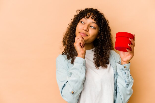 Joven afroamericana afro sosteniendo un regalo de San Valentín mirando hacia los lados con expresión dudosa y escéptica.