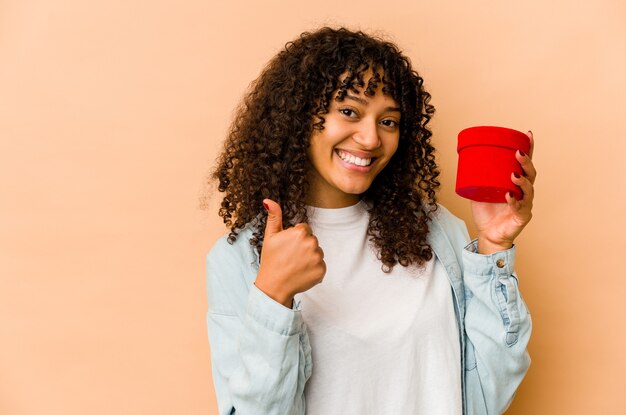 Joven afroamericana afro sosteniendo un regalo de San Valentín sonriendo y levantando el pulgar hacia arriba