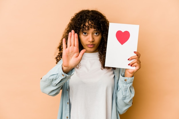 Joven afroamericana afro sosteniendo una tarjeta del día de San Valentín de pie con la mano extendida mostrando la señal de stop, impidiéndote.