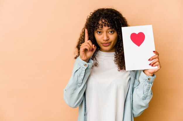 Joven afroamericana afro sosteniendo una tarjeta del día de San Valentín que muestra el número uno con el dedo.