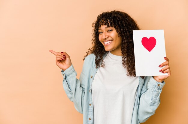 Joven afroamericana afro sosteniendo una tarjeta del día de San Valentín sonriendo y apuntando a un lado, mostrando algo en el espacio en blanco.