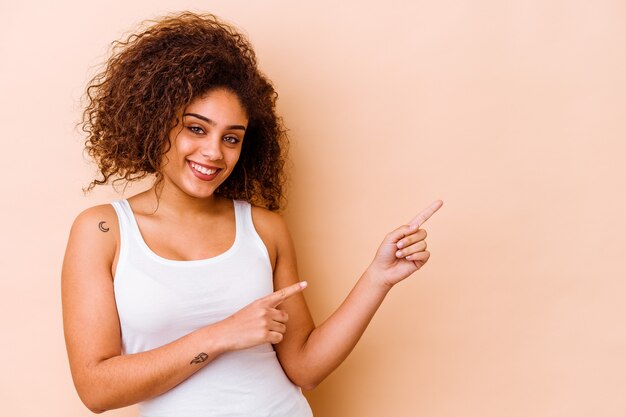 Joven afroamericana aislada en beige sonriendo alegremente apuntando con el dedo índice.