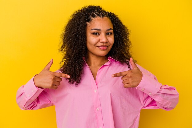 Joven afroamericana aislada en la pared amarilla persona apuntando con la mano a un espacio de copia de camisa, orgulloso y seguro