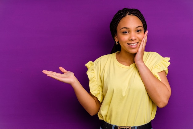 Joven afroamericana aislada en la pared amarilla tiene espacio de copia en una palma, mantenga la mano sobre la mejilla