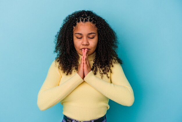 Joven afroamericana aislada en la pared azul rezando, mostrando devoción, persona religiosa en busca de inspiración divina