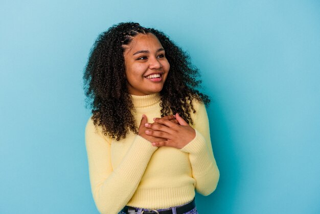 Joven afroamericana aislada en la pared azul riendo manteniendo las manos en el corazón