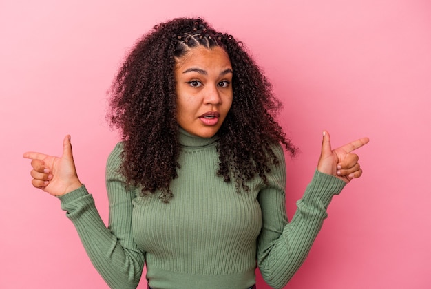 Joven afroamericana aislada en pared rosa apuntando a diferentes espacios de copia, eligiendo uno de ellos, mostrando con el dedo