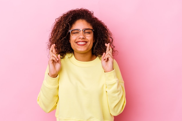 Joven afroamericana aislada en la pared rosa cruzando los dedos para tener suerte