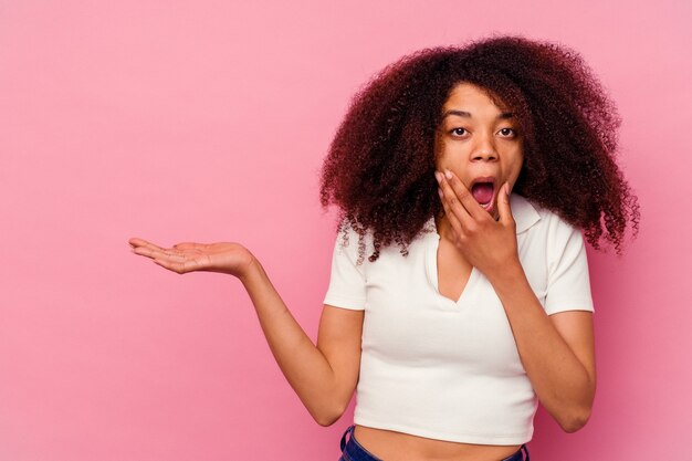 Joven afroamericana aislada en la pared rosa tiene espacio de copia en una palma, mantenga la mano sobre la mejilla. Asombrado y encantado.