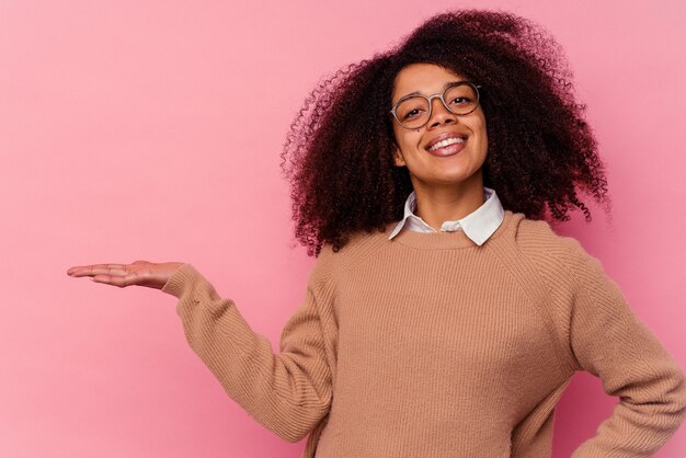 Joven afroamericana aislada en rosa mostrando un espacio de copia en una palma y sosteniendo otra mano en la cintura.