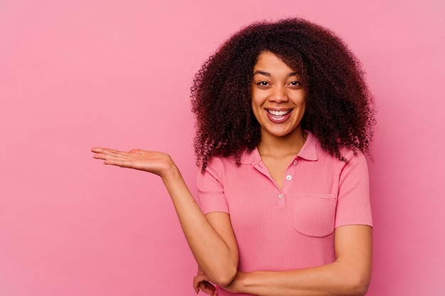 Joven afroamericana aislada en rosa mostrando un espacio de copia en una palma y sosteniendo otra mano en la cintura.