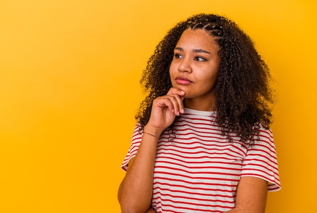 Joven afroamericana aislada sobre fondo amarillo mirando hacia los lados con expresión dudosa y escéptica.