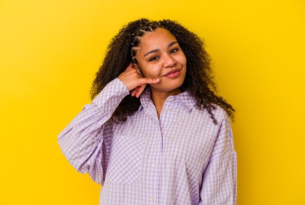 Joven afroamericana aislada sobre fondo amarillo mostrando un gesto de llamada de teléfono móvil con los dedos.