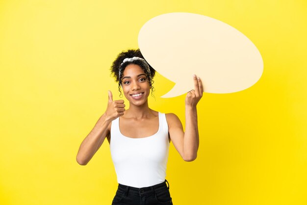 Joven afroamericana aislada sobre fondo amarillo sosteniendo un bocadillo vacío con el pulgar hacia arriba