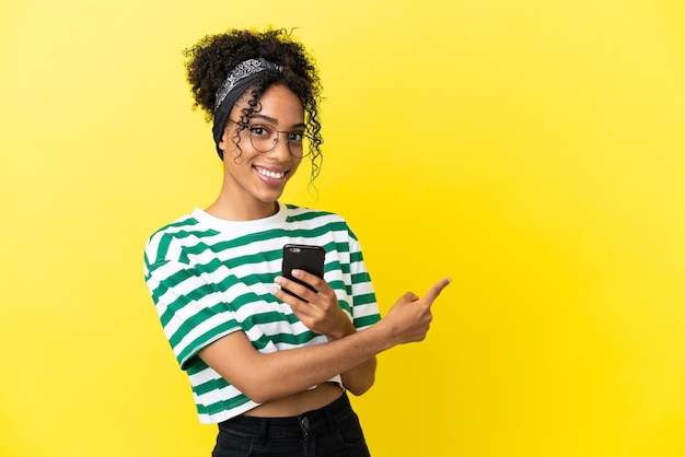 Joven afroamericana aislada sobre fondo amarillo con teléfono móvil y apuntando hacia atrás