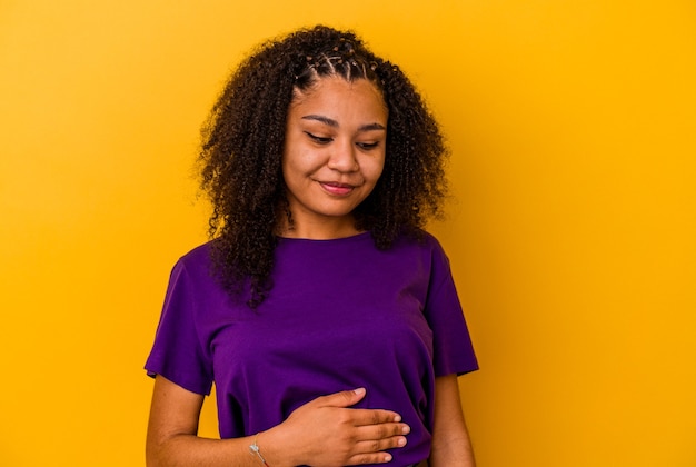 Joven afroamericana aislada sobre fondo amarillo toca la barriga, sonríe suavemente, comiendo y concepto de satisfacción.