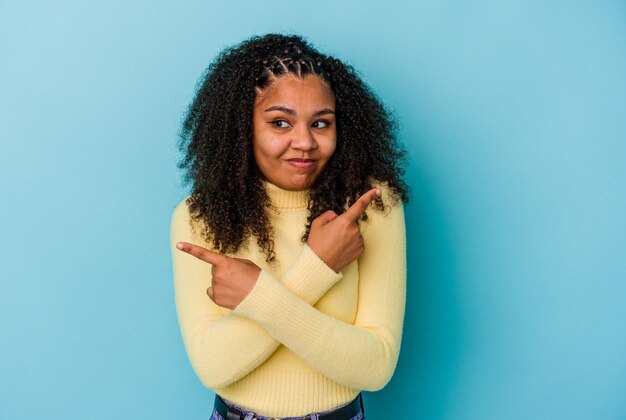 Joven afroamericana aislada sobre fondo azul apunta hacia los lados, está tratando de elegir entre dos opciones.