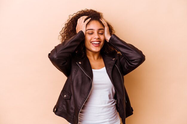 Joven afroamericana aislada sobre fondo beige se ríe con alegría manteniendo las manos en la cabeza. Concepto de felicidad.