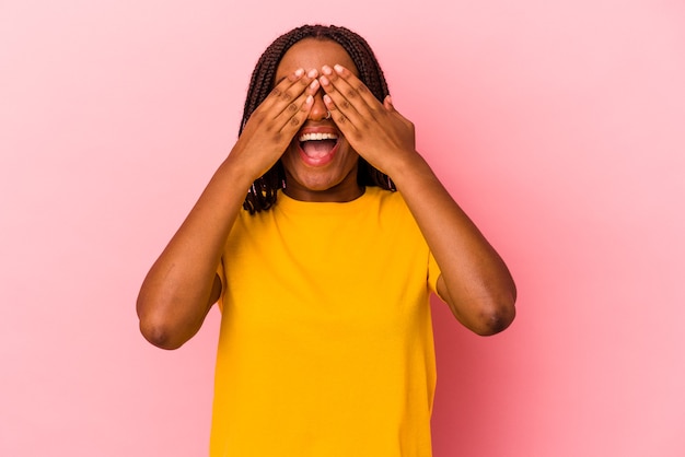 Joven afroamericana aislada sobre fondo rosa cubre los ojos con las manos, sonríe ampliamente esperando una sorpresa.