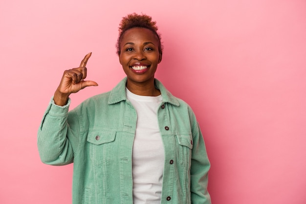 Joven afroamericana aislada sobre fondo rosa sosteniendo algo poco con los dedos índices, sonriendo y confiado.