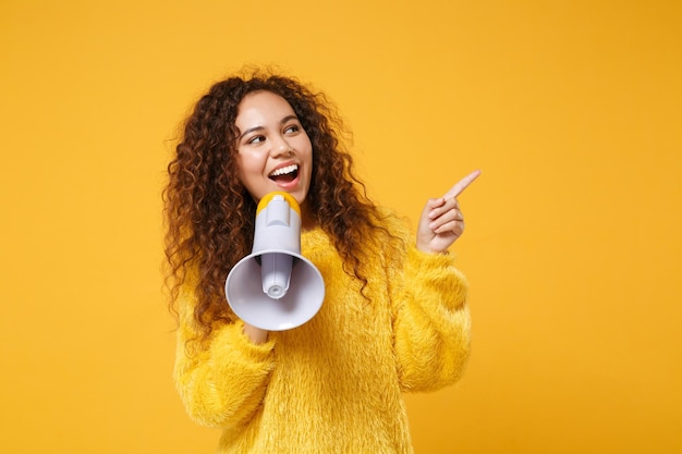 Una joven afroamericana alegre con un suéter de piel posando aislada en un fondo amarillo anaranjado de la pared. Concepto de estilo de vida de las personas. Simulacros de espacio de copia. Gritar en megáfono, señalando con el dedo índice a un lado.