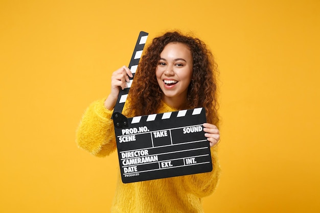 Una joven afroamericana alegre con suéter de piel posando aislada en un fondo naranja amarillo en el estudio. Concepto de estilo de vida de las personas. Simulacros de espacio de copia. Sosteniendo claqueta de cine negro clásico.