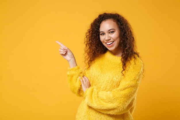 Una joven afroamericana alegre con suéter de piel posando aislada en un fondo naranja amarillo en el estudio. Gente emociones sinceras concepto de estilo de vida. Simulacros de espacio de copia. Señalando con el dedo índice a un lado.