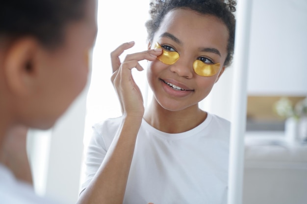 Joven afroamericana aplicando parches hidratantes debajo de los ojos mira el espejo