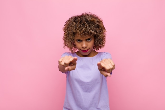 Joven afroamericana apuntando hacia adelante con ambos dedos y expresión enojada, diciéndole que cumpla con su deber en la pared rosa