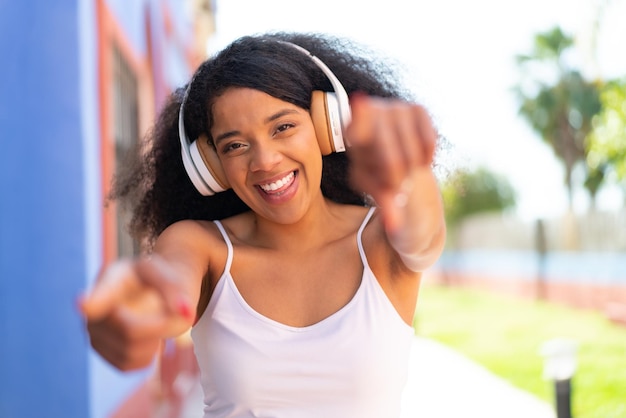 Una joven afroamericana con auriculares al aire libre te señala con el dedo mientras sonríe