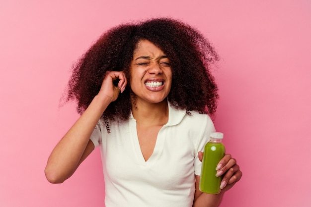 Joven afroamericana bebiendo un batido saludable aislado sobre fondo rosa cubriendo las orejas con las manos.