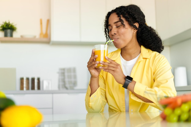 Joven afroamericana bebiendo jugo de naranja o mango fresco de vidrio sentado en el espacio de copia de la cocina