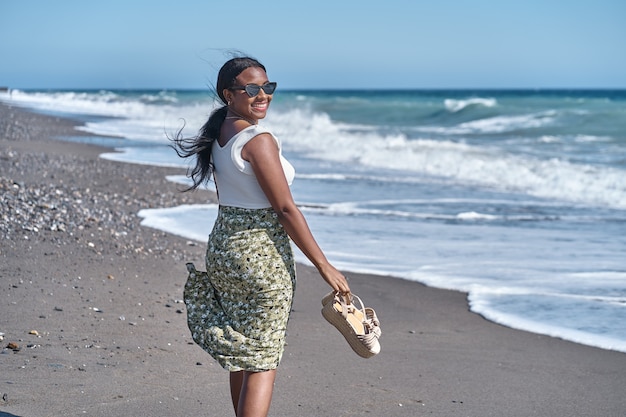 Joven afroamericana caminando por la orilla de la playa con sandalias en mano