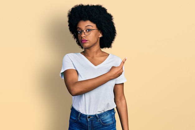 Joven afroamericana con camiseta blanca casual apuntando con el dedo de la mano hacia un lado mostrando publicidad, rostro serio y tranquilo