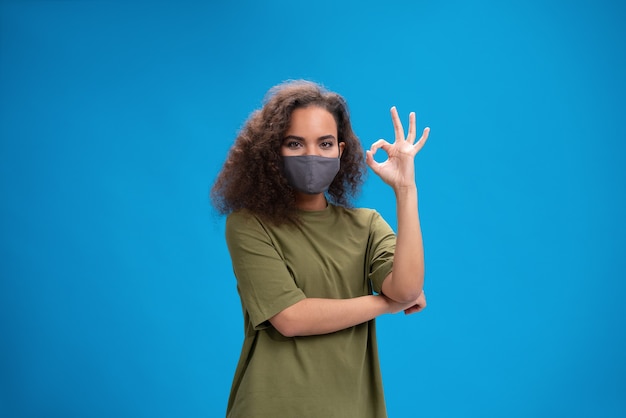 Joven afroamericana en camiseta caqui en mascarilla reutilizable mostrando gesto OK