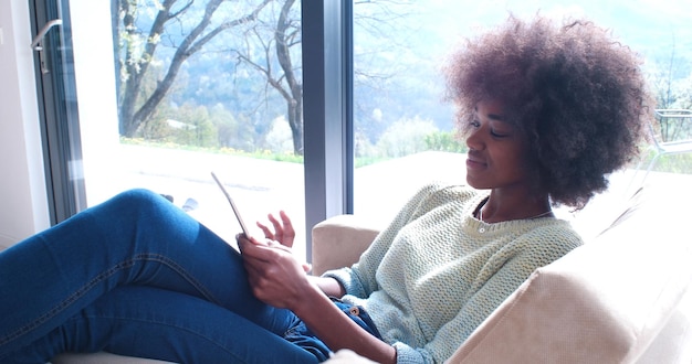 Joven afroamericana en casa relajándose en su lujosa sala de estar leyendo una tableta digital, navegar por Internet y trabajar