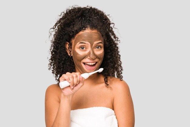 Joven afroamericana cepillando los dientes y aplicándose una máscara facial aislada