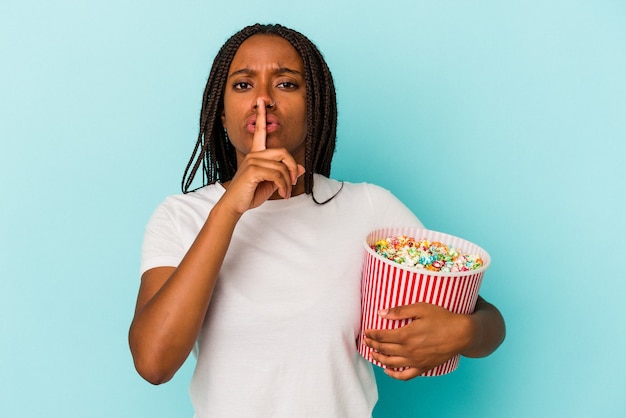 Joven afroamericana comiendo palomitas de maíz aislado sobre fondo azul manteniendo un secreto o pidiendo silencio.