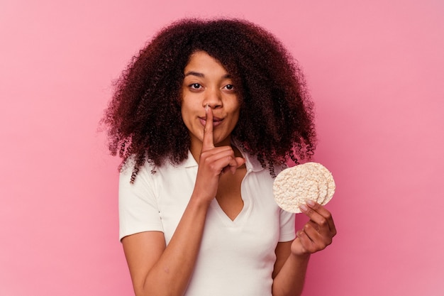 Joven afroamericana comiendo un pastel de arroz aislado en rosa guardando un secreto o pidiendo silencio.