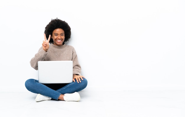 Joven afroamericana con una computadora portátil sentada en el suelo sonriendo y mostrando el signo de la victoria