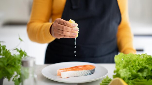 Joven afroamericana en delantal vierte jugo de limón cocina pescado en la mesa con verduras frescas