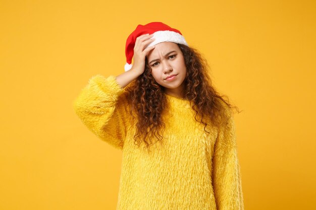 Una joven afroamericana disgustada con suéter de piel, sombrero de Navidad aislado de fondo amarillo. Feliz Año Nuevo 2020 concepto de vacaciones de celebración. Simulacros de espacio de copia. Ponerse la mano en la cabeza.