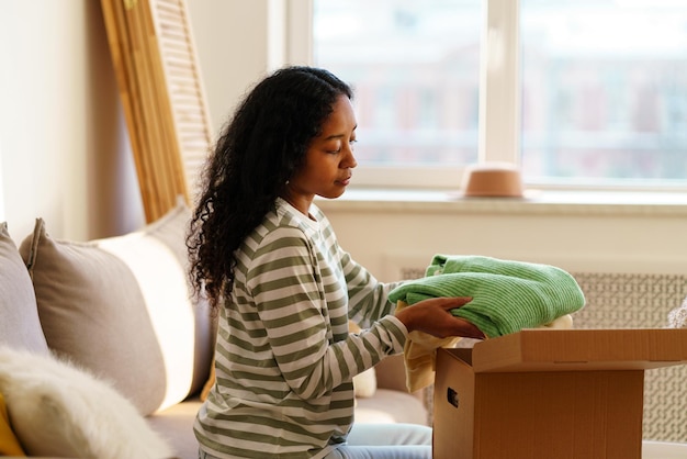 Joven afroamericana empacando cosas en una caja de cartón mudándose y regalando ropa