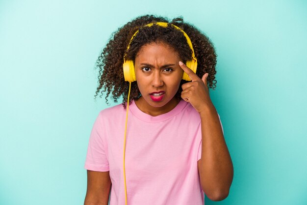 Joven afroamericana escuchando música aislada sobre fondo azul mostrando un gesto de decepción con el dedo índice.